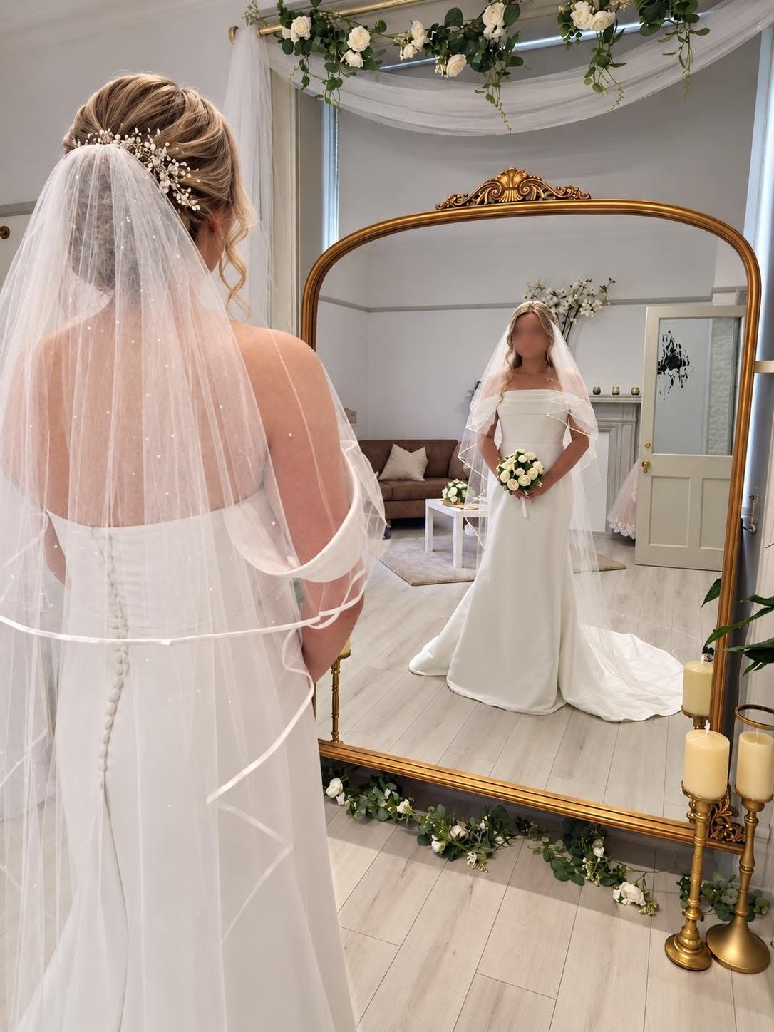A bride in a satin off-the-shoulder gown and long veil looking into the boutique’s gold mirror