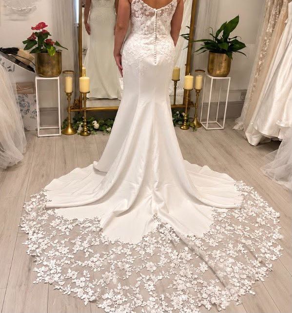A bride in a fitted lace-backed gown with a long floral lace train, in front of the boutique’s gold mirror